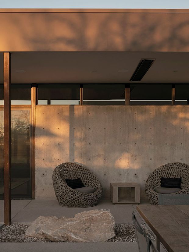 A warm, moody architectural exterior or covered outdoor sitting area with exposed concrete walls, dark timber or metal framing, narrow clerestory glazing, and a flat overhanging roof. Two woven lounge chairs and a small concrete side table sit on large-format paving beside a gravel bed with a natural stone feature. Strong late-afternoon shadows create an earthy, refined atmosphere.