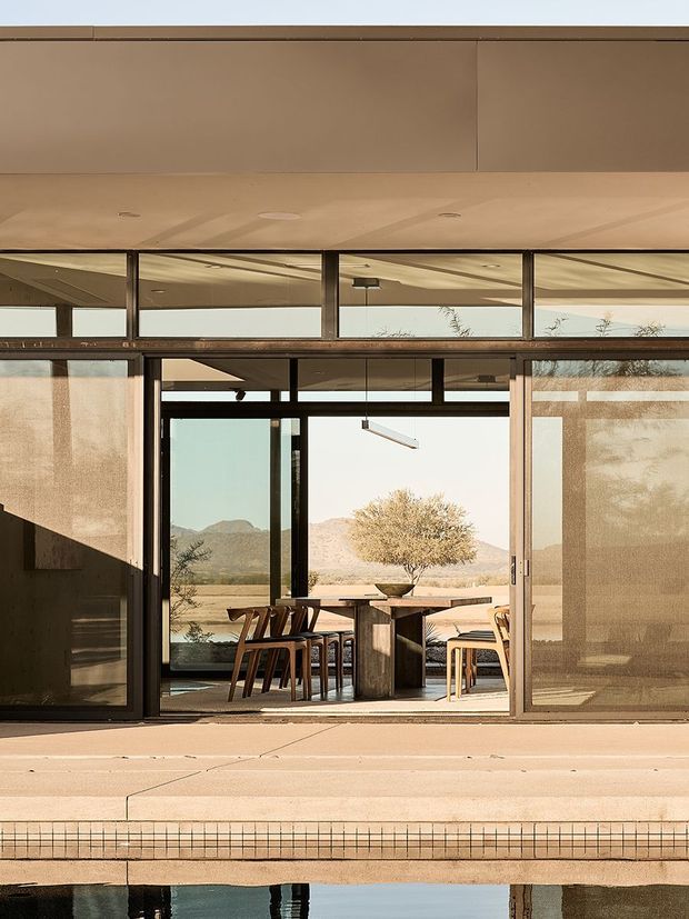 A warm, minimalist modern pavilion viewed from a poolside terrace, with broad floor-to-ceiling glazing, dark slender window and door frames, a deep flat projecting roof, and integrated roller screens. Inside is a dining area with a timber table and chairs, opening toward a dry mountainous landscape and a planted courtyard-like setting. The foreground includes a tiled pool edge and reflective water.