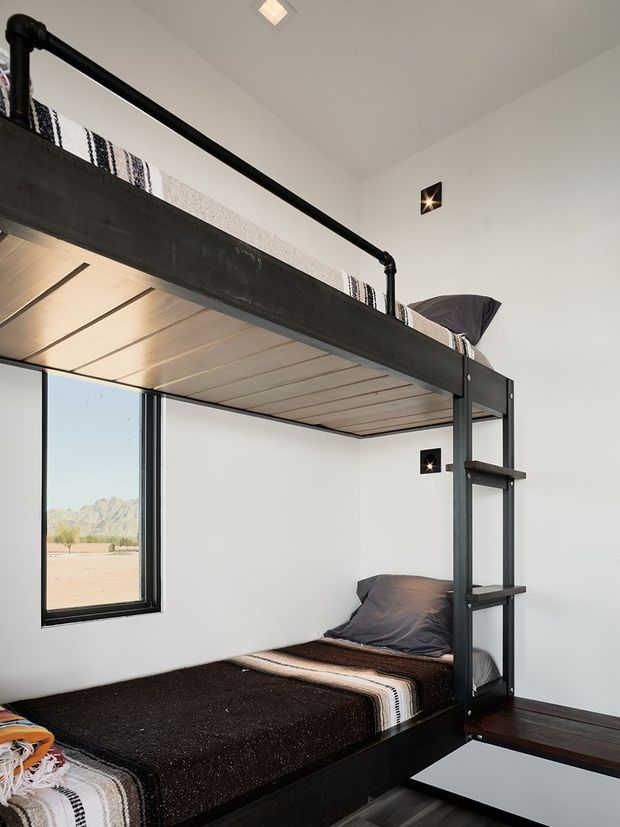 Compact bunk bedroom with a black metal-framed loft bunk bed, dark bedding, integrated ladder shelving, and warm timber slatted undersurface. White walls and black-framed windows create a clean modern interior, with a desert mountain landscape visible outside.