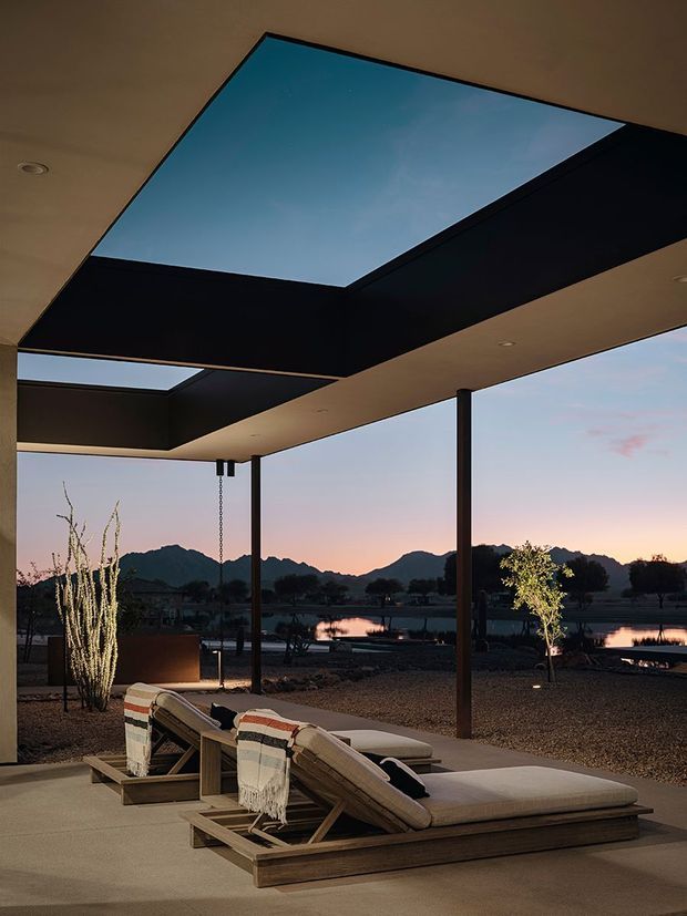A covered outdoor lounge terrace at dusk, furnished with two low timber-framed chaise lounges and neutral cushions. Slim dark structural posts support a broad flat roof with large rectangular skylight openings, creating a dramatic geometric ceiling. The open sides frame a calm water feature or pool and distant mountain silhouettes, with sparse desert-style landscaping and warm recessed lighting.