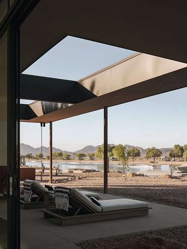 A dramatic contemporary outdoor living area viewed from inside through fully open glazing, featuring a shaded concrete terrace, two reclining lounge chairs, slim dark structural posts, and a large angular roof canopy with skylight-like openings. Beyond the terrace is a sparse landscaped site with water and rugged mountains in the distance.