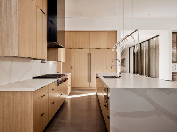 A contemporary galley-style kitchen with light timber veneer cabinetry, white stone benchtops, a large waterfall-edge island, integrated appliances, a dark polished concrete floor, and sculptural loop pendant lighting. A glazed internal partition or window runs along the right side, allowing light into the space.