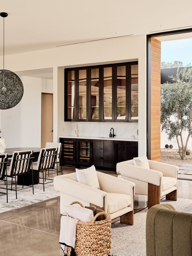 A bright, open-plan contemporary interior combining a living area, dining area, and compact kitchen/bar zone. The space features pale plaster walls and ceiling, large floor-to-ceiling glazing, dark timber-framed internal windows, dark built-in cabinetry, a light stone benchtop, polished concrete flooring, and neutral upholstered seating. A sculptural dark pendant hangs over the dining table, while an outdoor courtyard with a tree is visible through the glazing.