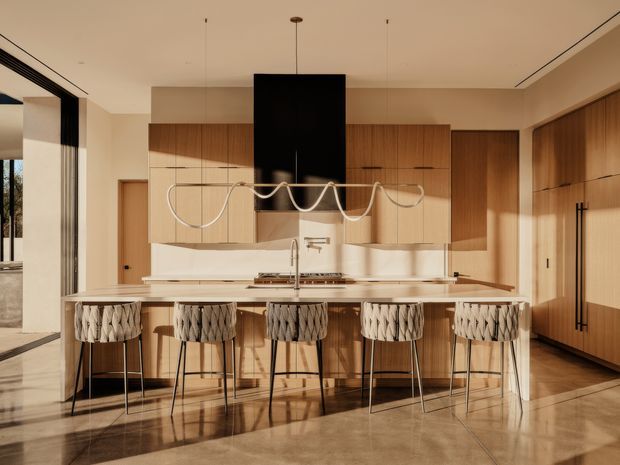 A sophisticated contemporary kitchen centered on a large island with a pale stone benchtop and multiple woven bar stools. Warm timber cabinetry extends across the back wall and into a tall storage bank, contrasting with cream walls, a black rangehood, and a sculptural linear pendant light. The space has polished stone or concrete-look flooring, integrated appliances, generous glazing, and strong afternoon sunlight.