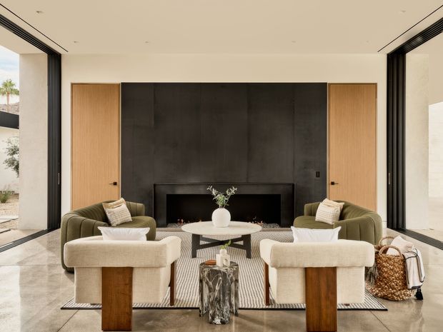 A refined contemporary living room arranged around a wide, dark fireplace feature wall. Two olive-green lounge chairs face the fireplace, with two cream boucle armchairs in the foreground, a low round coffee table, and a sculptural side table. Full-height black-framed glazing opens the room to an outdoor courtyard, creating a bright indoor-outdoor connection. The space has pale stone flooring, warm timber wall panels and doors, a light ceiling, and a restrained neutral palette.