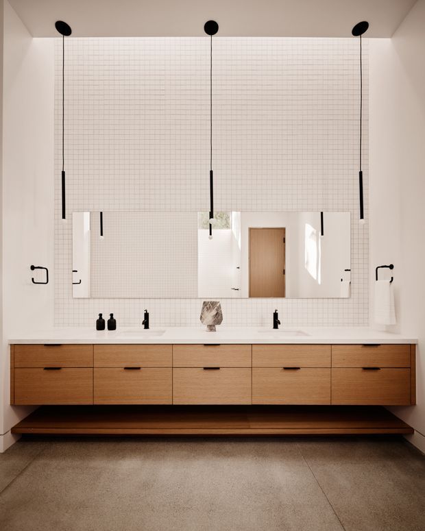 A refined contemporary bathroom with a long floating timber vanity, twin integrated sinks, black wall-mounted fixtures, and a large mirrored cabinet. The walls are finished in small white square tiles, complemented by minimalist black pendant lights, black hardware, and a warm wood door. A broad concrete or stone-look floor adds a restrained, tactile finish.