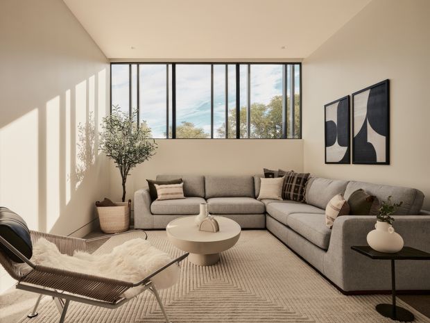 A bright, refined living room with a large grey sectional sofa, neutral textured rug, sculptural lounge chair, side table, potted tree, and abstract monochrome artwork. Floor-to-ceiling black-framed glazing brings in abundant daylight and views of trees, while the high sloped ceiling creates an airy contemporary feel.