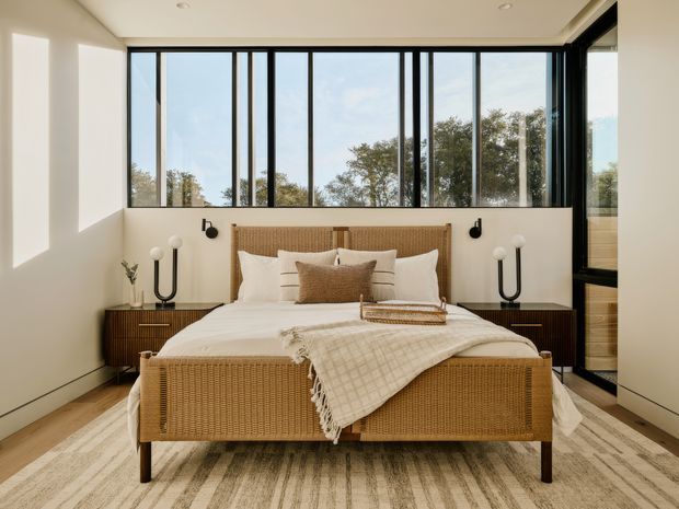 A bright, contemporary bedroom centred around a woven rattan or cane bed frame with crisp white bedding and neutral textured throws. Matching dark wood bedside cabinets, sculptural table lamps, and black wall sconces flank the bed. Full-height black-framed windows provide abundant natural light and views of trees, while a large neutral striped rug spans the timber floor. The room has a clean, warm, refined feel with a low ceiling and simple recessed lighting.