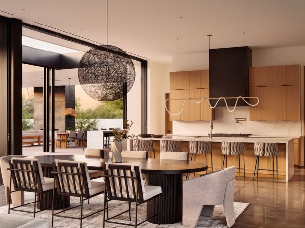 Open-plan contemporary kitchen and dining area with a long island, timber cabinetry, dark feature cabinetry, integrated appliances, and sculptural pendant lighting. A large dining table with upholstered and timber-framed chairs occupies the foreground, while floor-to-ceiling glazing opens toward an outdoor area. The palette is warm and refined, with timber, cream surfaces, dark accents, and polished flooring.