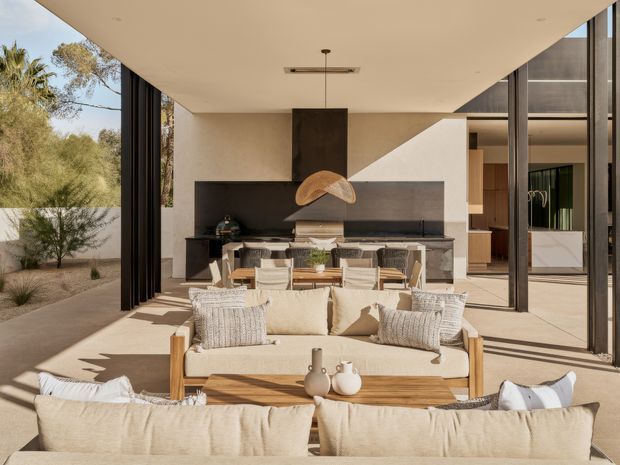 A sunlit, open-plan indoor-outdoor living and dining space with extensive floor-to-ceiling glazing and fully opened dark-framed sliding doors. A large neutral upholstered sofa faces a timber dining table and chairs, with a built-in dark stone or concrete kitchen counter and cabinetry beyond. The room opens directly to a landscaped arid garden, and a sculptural pendant light hangs over the dining area. The design is clean-lined and contemporary, with warm natural textures and strong indoor-outdoor connection.