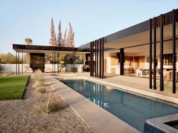 A single-storey modern residence arranged around a long rectangular swimming pool. The open-plan living area has broad floor-to-ceiling glazing and extensive sliding glass doors framed by dark vertical metal elements. Covered outdoor living and dining areas extend beneath flat roof planes, with a minimalist landscaped courtyard featuring gravel, ornamental grasses and lawn. Warm timber and neutral finishes are visible inside, while the pool forms the central focus of the composition.