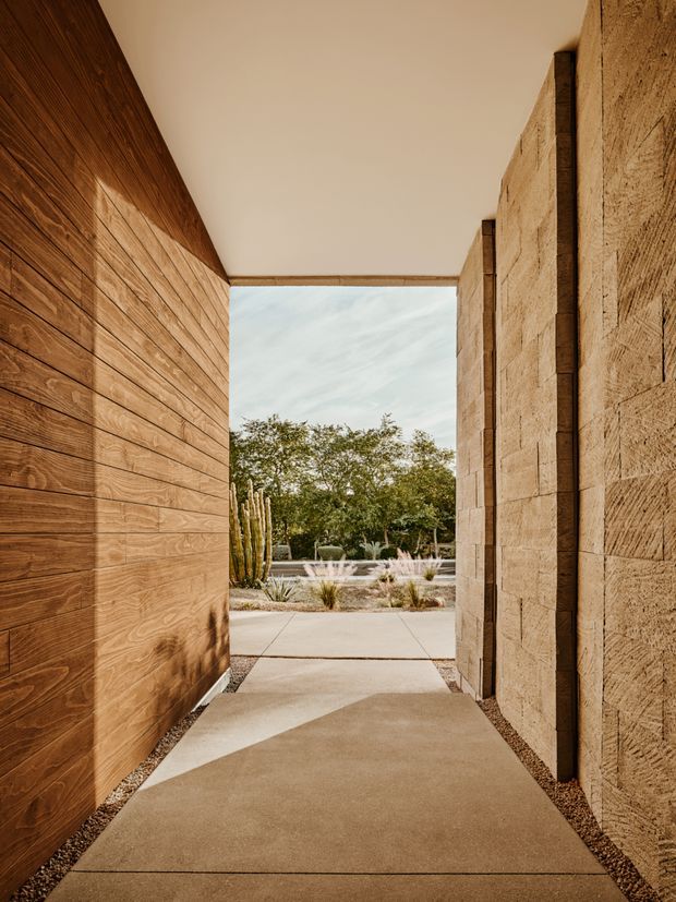 A covered architectural entry passage leading toward a landscaped courtyard. The narrow walkway is framed by warm horizontal timber-clad walls on the left and textured tan masonry walls with dark vertical timber or metal accents on the right. A smooth pale ceiling and concrete paving create a minimalist, linear composition, opening to gravel landscaping, cacti, ornamental grasses, trees, and a shallow water feature beyond.