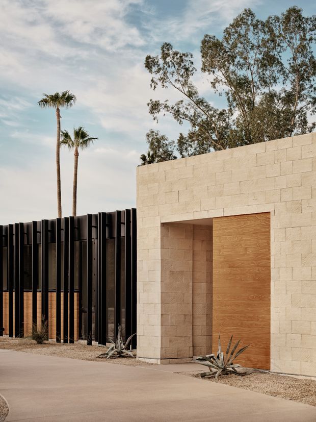 Exterior architectural view of a minimalist compound entrance, featuring a pale rectangular masonry volume with a large flush timber door, set beside a dark vertical slatted screen. Desert-style gravel landscaping, agave plants, and tall palms and eucalyptus trees frame the warm modern facade.