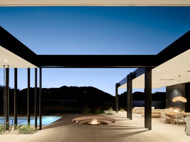 Contemporary open-plan residence with dramatic black steel framing and broad flat roof planes around a large open-air courtyard. The space features a smooth concrete terrace, integrated circular fire pit seating, a swimming pool, and a warmly lit dining and living area opening directly onto the outdoor space. Dark mountain silhouettes are visible beyond the property under a clear blue sky.