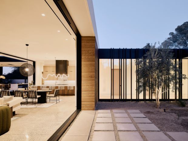 Contemporary residential courtyard view through expansive floor-to-ceiling glazing, showing a refined open-plan living and dining area connected to a paved outdoor courtyard. The interior features pale stone or terrazzo flooring, warm timber wall cladding, black-framed glazing, recessed lighting, and a modern dining and kitchen zone. Outside, large square paving slabs are set into a dark gravel landscape, with a black architectural screen and slender trees creating privacy.