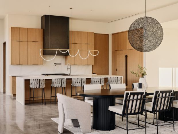 Open-plan contemporary kitchen and dining area with warm timber cabinetry, a large dark rangehood, white island bench with bar seating, and a black dining table surrounded by modern upholstered and framed chairs. A sculptural pendant light hangs over the dining table, while polished concrete-style flooring and a neutral palette create a refined, spacious interior.