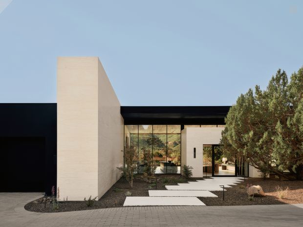 Contemporary single-storey residence with a minimalist geometric facade, composed of white rendered masonry volumes, a black horizontal wing, and extensive floor-to-ceiling glazing. A landscaped entry courtyard with stepping slabs, gravel, mature planting, and a paved driveway leads to the recessed front entrance. The composition has a strong indoor-outdoor connection and clean rectilinear lines.