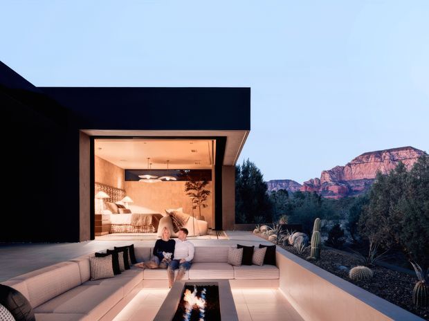 A dramatic modern desert residence at dusk, with a deep covered outdoor living terrace opening directly to an illuminated interior bedroom or lounge. A large built-in pale sectional surrounds a rectangular fire pit, while dark minimalist walls frame expansive glazing and a landscaped garden. Red rock cliffs and mature vegetation form the distant backdrop.