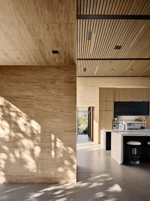 A warm, high-end contemporary kitchen viewed from an open-plan interior. Large-format beige stone or porcelain surfaces clad the walls and floor, while a slatted timber ceiling with dark structural bands adds texture and visual rhythm. The kitchen features tall timber cabinetry, a dark integrated rangehood or backsplash zone, a pale island with a waterfall edge, and dark fluted panel detailing with a round stool. Natural sunlight creates dappled shadows across the stone surfaces, and a glazed opening provides a view toward a mountainous landscape.