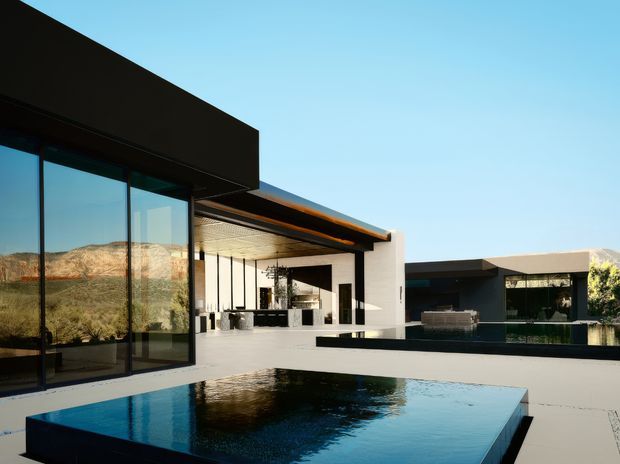 A contemporary single-storey residence arranged around a reflective rectangular pool. The home features strong horizontal and rectilinear volumes, extensive floor-to-ceiling glazing, dark flat roof planes, white rendered walls, and a covered outdoor entertaining area with a timber-lined ceiling. A dry mountainous landscape is visible beyond the property.