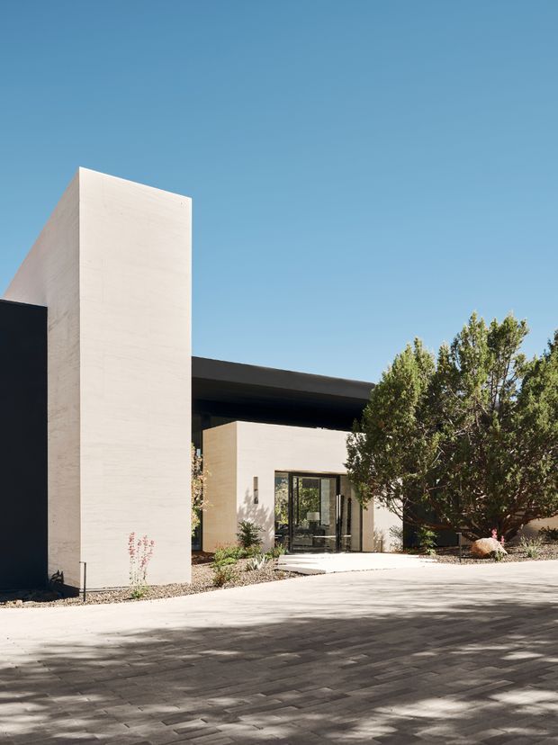 Exterior view of a minimalist contemporary residence with strong geometric forms, pale rendered walls, a dark flat roof canopy, and large glazed entry doors. The building is approached by a broad paved driveway or forecourt, with drought-tolerant landscaping and mature trees around the entrance under a clear blue sky.