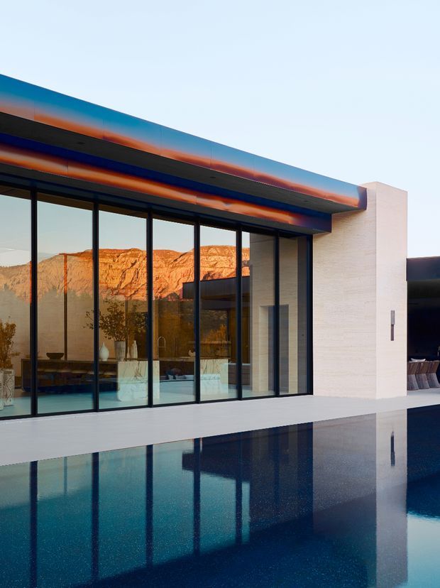 A contemporary flat-roofed building exterior beside a reflecting swimming pool, featuring extensive floor-to-ceiling glazing with dark slender frames, white masonry walls, and a vivid blue-and-orange roof-edge finish. The glazing reflects a dramatic red-rock mountain landscape, while interior furnishings and a tree are visible behind the glass.