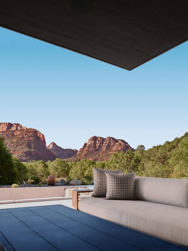 An outdoor covered terrace with a built-in light upholstered sofa and patterned cushions, overlooking a dramatic red-rock mountain landscape. The space features dark wood soffit overhead, a dark tiled or slab floor, a low pale stucco parapet, and sparse desert landscaping. The composition feels contemporary, calm, and strongly connected to the surrounding natural setting.