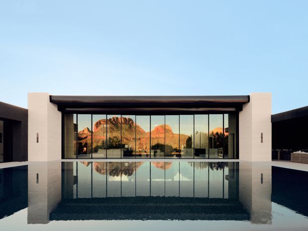 A symmetrical modern building facade viewed across a reflecting pool. The structure has a low, rectilinear form with dark side walls, pale rendered vertical piers, a deep dark roof or canopy edge, and a broad expanse of floor-to-ceiling glazing. The glass reflects a dramatic rocky desert landscape and the pool mirrors the entire facade. The scene is clean, minimalist, and strongly geometric under a clear blue sky.