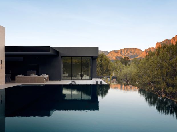 A contemporary dark-clad residence overlooking a reflective swimming pool, with broad floor-to-ceiling glazing opening toward a rugged sunlit mountain landscape. The architecture has strong rectilinear volumes, a flat roof, deep black exterior surfaces, and an integrated pool terrace. The scene is photographed at calm golden-hour light.