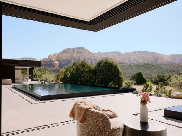 A refined outdoor terrace framed by a deep contemporary roof overhang, overlooking a rectangular infinity-edge pool and a dramatic rugged mountain landscape. The pale tiled or concrete terrace includes a curved textured lounge chair, round dark side table, sculptural vase with flowers, and a partially visible outdoor seating area. The scene is open, sunny, and strongly connected to the surrounding natural setting.