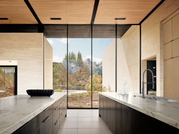 A spacious contemporary kitchen with two parallel marble-topped islands or counters, dark wood cabinetry, pale stone walls, and a slatted timber ceiling with black structural accents. Full-height glazing opens views toward trees and rugged mountains, bringing abundant natural light into the refined, minimalist interior.