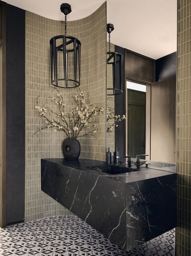 A sophisticated bathroom with a wall-mounted double vanity clad in dark veined stone, black fixtures, and a large mirrored wall. Curved walls are finished in narrow vertically arranged muted green-grey tiles, while the floor features a bold geometric black, white, and grey patterned tile. Black cage pendant lights and a sculptural vase with flowering branches add decorative detail. The space has a dramatic, refined, art deco-inspired character.