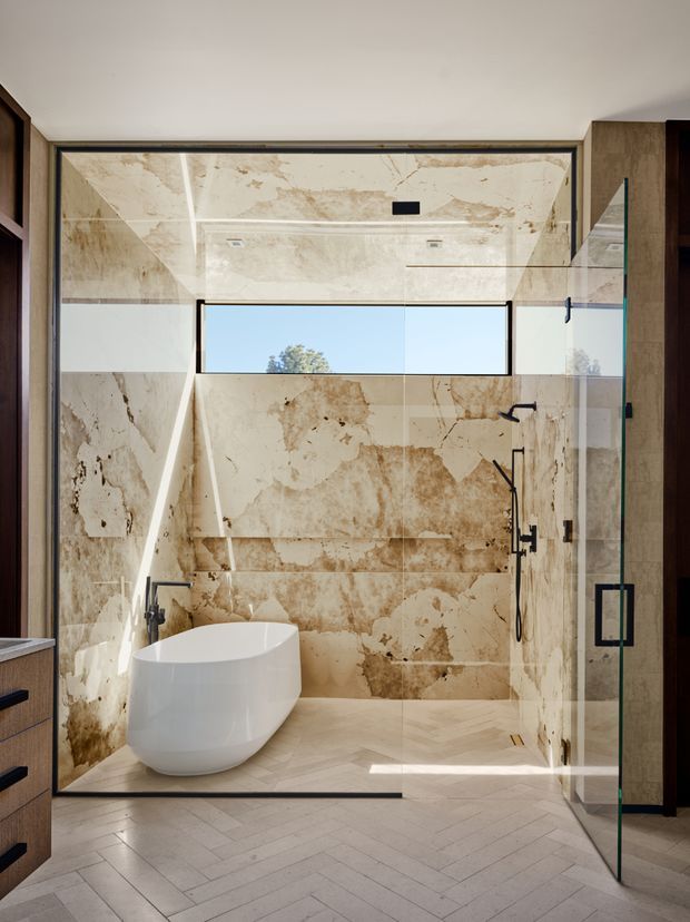 A luxurious contemporary bathroom featuring a freestanding white bathtub, a large walk-in shower enclosed by frameless glass, black fixtures, and extensive warm beige veined stone wall and floor finishes. A narrow horizontal window provides natural light, while dark timber-toned joinery frames the room.