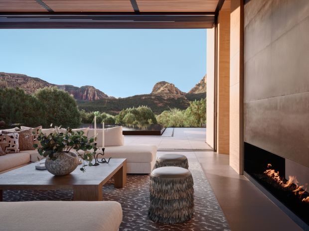 A refined contemporary living room opens directly onto a spacious terrace through fully retractable glazing or an open wall. The room features a large pale sectional sofa, low timber coffee table, patterned area rug, sculptural fringed ottomans, and a dramatic floor-to-ceiling fireplace clad in smooth warm-toned stone or concrete. A timber slatted overhead screen shades the opening, while landscaped planting and rugged red rock formations are visible beyond.