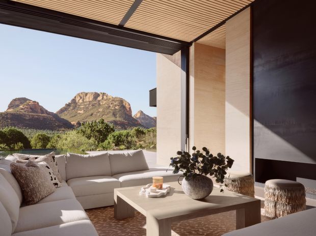 A refined contemporary outdoor living room opens broadly toward a dramatic red-rock mountain landscape. The space features a large pale sectional sofa, low rectangular coffee table, sculptural vase and plant, textured ottoman, dark vertical fireplace or feature wall, pale stone-toned walls, and a slatted timber ceiling with dark structural framing. The indoor-outdoor setting is bright, warm, and strongly connected to the surrounding landscape.