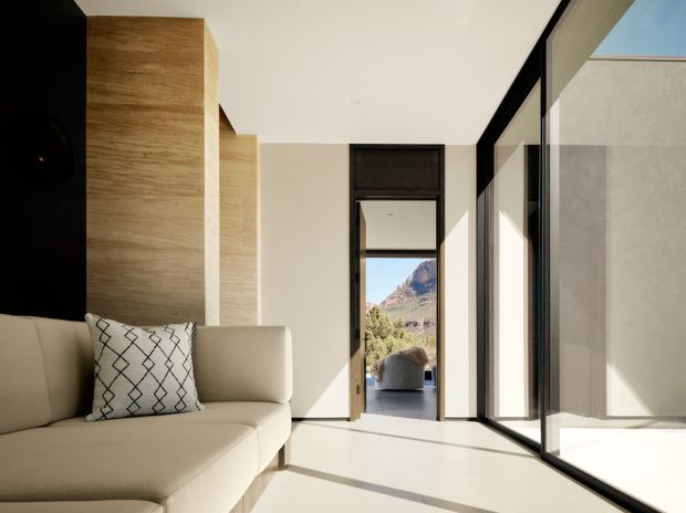 Bright contemporary interior hallway or living area with a large cream sectional sofa in the foreground, warm wood wall paneling, white walls and flooring, and expansive black-framed floor-to-ceiling glazing along the right side. A doorway at the rear opens toward an outdoor seating area and mountainous landscape. The space has clean lines, strong natural light, and a minimal, refined character.