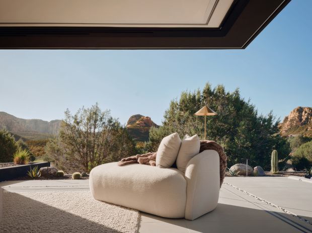 A contemporary outdoor lounge area framed beneath a broad roof overhang, featuring a large cream upholstered lounge chair with cushions and a textured rug on a pale paved terrace. Desert landscaping with sculptural shrubs, cacti, and red-rock mountains is visible beyond, creating a relaxed resort-like setting.