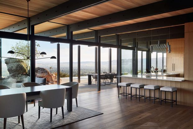 A spacious open-plan modern interior combining a kitchen, dining area and living zone. The room features extensive floor-to-ceiling glazing with expansive elevated landscape views, dark exposed steel beams and mullions, warm timber ceiling boards and timber flooring, and a large timber kitchen island with integrated seating. Sculptural pendant lighting and restrained contemporary furnishings create a refined connection to the outdoors.