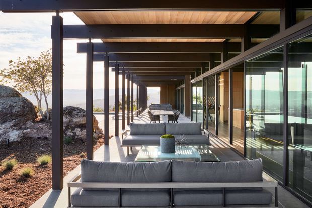 A long, covered outdoor terrace beside a modern glass-walled building, furnished with grey sofas, coffee tables, and dining seating. Dark structural posts support a timber-lined ceiling, while the terrace overlooks a rocky landscaped setting and distant water or landscape views. The composition emphasizes the deep linear veranda, indoor-outdoor connection, and expansive glazing.
