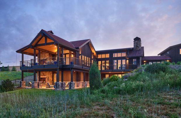 A large multi-level mountain-style residence photographed at dusk, set within natural grasses and landscaped vegetation. The home features dark timber and stone exterior finishes, multiple pitched gables, broad covered balconies, extensive glazing, warm interior lighting, and a prominent stone chimney.