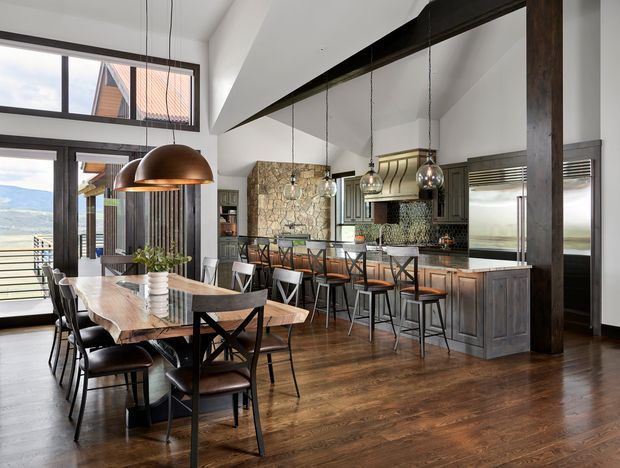 Open-plan kitchen and dining interior with a high vaulted ceiling, exposed dark structural beams, extensive glazing, and views toward a mountainous landscape. A large timber dining table sits beside a long kitchen island with bar seating. The kitchen features dark timber cabinetry, a patterned tile backsplash, stainless-steel appliances, stone surfaces, and pendant lighting. The overall aesthetic is warm, rustic-modern, and lodge-like.