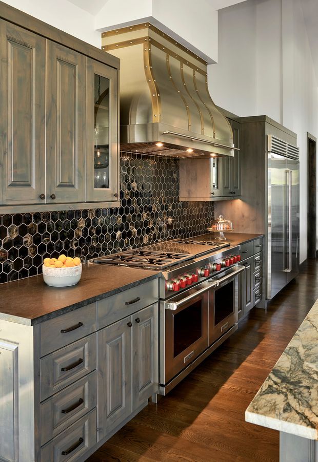 A detailed high-end kitchen with grey distressed-style fixed cabinetry, dark natural stone benchtops, a large professional stainless-steel range cooker, and a sculptural metallic rangehood with brass-toned trim. The backsplash features glossy dark hexagonal tiles with light grout. Dark timber flooring, stainless appliances, and warm under-cabinet lighting create a dramatic, richly layered kitchen interior.