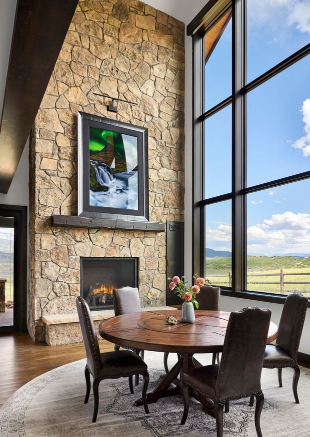 A rustic dining area with a round dark timber table and upholstered dining chairs positioned beside a large stone fireplace. The fireplace wall is built from irregular natural stone with a timber mantel and framed artwork above, while expansive black-framed windows provide views of an open rural landscape. The room has timber flooring, a large patterned rug, and abundant natural light with a warm, lodge-inspired atmosphere.