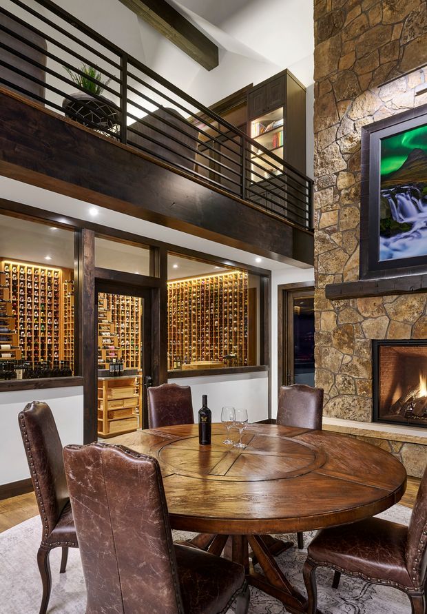 A warm, double-height dining area with a large round timber dining table and brown leather dining chairs in the foreground. Behind it are extensive illuminated built-in wine storage racks, a stone-clad fireplace with a mounted screen, and a dark timber mezzanine with black metal railings and integrated shelving. The space has a rustic, lodge-inspired character with dramatic layered lighting and rich natural materials.