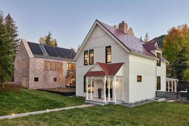 Exterior view of a renovated or newly built residential property set in a leafy suburban or wooded setting. The main house is a two-storey white weatherboard dwelling with a steep gabled roof, decorative gable detailing, red metal roof accents, brick chimney, covered front porch, and dark-framed windows. A modern adjoining brick structure with extensive solar panels is visible to the left, connected by glazed elements. Paved paths, lawn, mature trees, and a dark perimeter fence complete the scene.