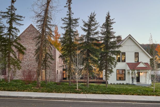 Exterior street view of a two-storey residential property comprising a white gable-fronted house with a covered entry porch and a larger adjoining brick-clad volume. Mature evergreen and deciduous trees partially screen the building, with a suburban sidewalk and road in the foreground. The architecture features steep pitched gable roofs, white horizontal siding, brick masonry, dark-framed windows, and a small red-toned metal porch roof.