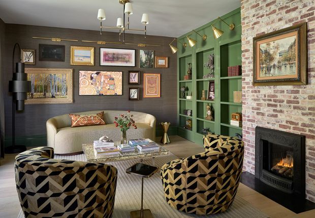 A richly layered living room with a cream sofa, two bold geometric-patterned lounge chairs, a low coffee table, and extensive framed artwork. The room combines a dark textured wall, deep green built-in shelving with decorative lighting, and an exposed brick fireplace wall with a black firebox. Brass and gold-toned lighting and accessories add warmth and visual contrast.