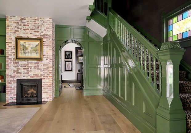 A traditional interior hallway and stairway centered on a richly detailed green-painted staircase with paneled wainscoting, turned balusters, and a substantial newel post. A stained-glass window lights the upper stair, while a whitewashed exposed-brick fireplace wall with framed artwork anchors the left side. Wide light timber floorboards, dark timber stair treads, built-in shelving, and an arched opening into a further room complete the composition.