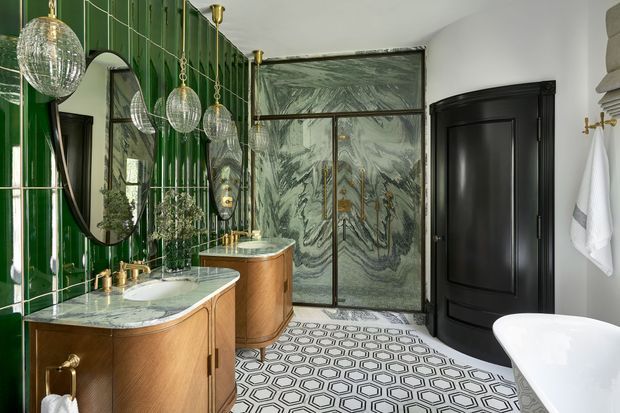 A luxurious, richly detailed bathroom featuring a double vanity with curved timber cabinetry and pale stone benchtops, brass tapware, a glossy emerald-green tiled feature wall, multiple mirrors, pendant globe lights, patterned geometric floor tiles, a marble-look glazed shower enclosure, and a freestanding white bathtub. Black panelled doors and trims add strong contrast.