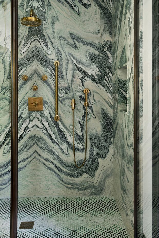 A close-up view of a luxurious walk-in shower enclosed by dark-framed glass. The walls are clad in dramatic green, grey, and white veined stone, complemented by antique brass shower fittings, controls, and hand shower. The floor features a geometric mosaic tile pattern in green, grey, black, and white. A partial reflection is visible in the glazing.